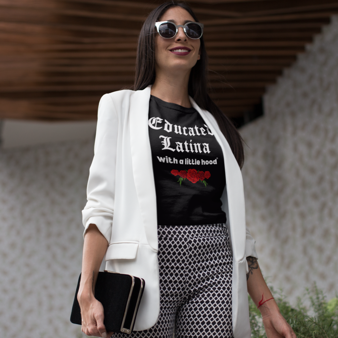 Woman wearing a black t-shirt with 'Educated Latina with a Little Hood' text and graphics, white blazer, and patterned pants outdoors.