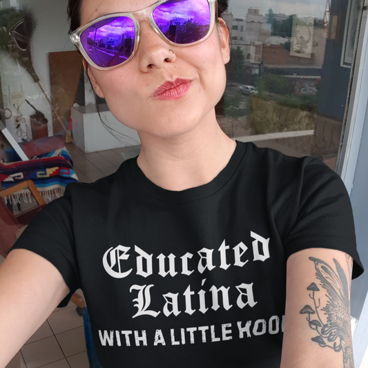 Woman wearing a black t-shirt with 'Educated Latina with a Little Hood' text, purple sunglasses, and tattoos.